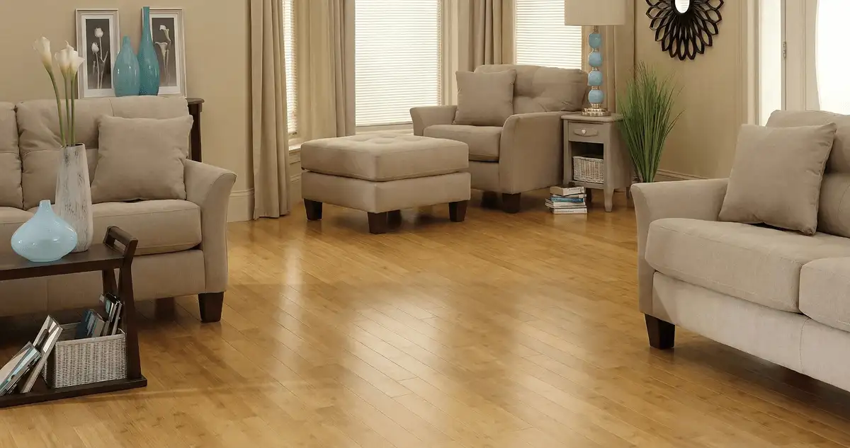 A living room with professionally installed bamboo flooring, beige sofas, a matching ottoman, a side table with a blue lamp, vases, framed pictures, and large windows with blinds and curtains in Henderson, NV